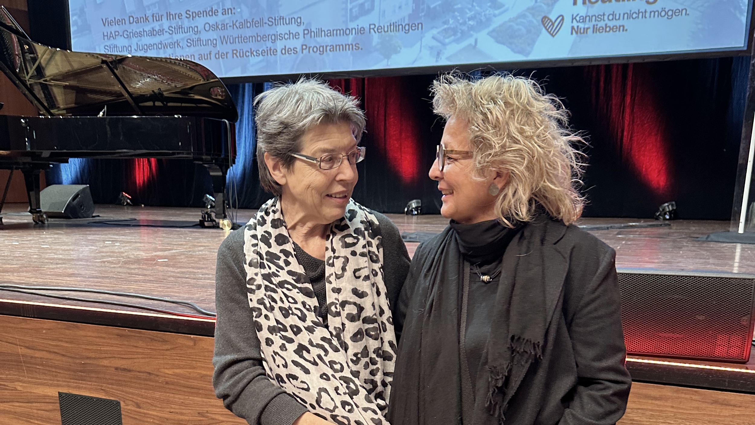 Susanne Stutzmann und Beate Müller-Gemmeke stehen auf der Bühne des Theaters Reutlingen und unterhalten sich lächelnd. Im Hintergrund sind Bühne und Konzertflügel zu sehen.