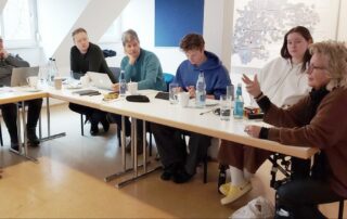 Beate Müller-Gemmeke sitzt an einem Tisch in einem hellen Raum und spricht engagiert zu einer Diskussionsrunde. Mehrere Menschen hören aufmerksam zu, vor ihnen stehen Wasserflaschen und Notizen. Die Szene zeigt eine konzentrierte politische Debatte.