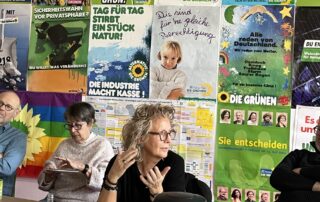 Diskussionsrunde der Grünen in Waiblingen zum Thema Bürgergeld und Grundsicherung. Beate Müller-Gemmeke spricht an einem Tisch, andere Teilnehmende hören aufmerksam zu. Im Hintergrund hängen historische Wahlplakate von Bündnis 90/Die Grünen.