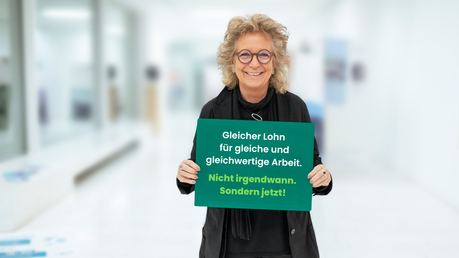 Beate Müller-Gemmeke steht vor hellem Hintergrund und lächelt in die Kamera. Sie hält ein grünes Schild mit der Aufschrift: „Gleicher Lohn für gleiche und gleichwertige Arbeit. Nicht irgendwann. Sondern jetzt!“ Sie trägt schwarze Kleidung und eine runde Brille.