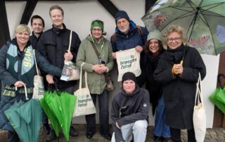 Eine Gruppe von Wahlkämpfer:innen steht dicht beieinander vor einer Hauswand. Es ist der Start zum Haustür-Wahlkampf. Einige halten Stofftaschen mit der Aufschrift „Stabil in bewegten Zeiten“. Zwei grüne Regenschirme lehnen am Boden. Die Stimmung wirkt trotz Regen freundlich und entschlossen.