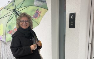 Beate Müller-Gemmeke steht bei Regen in Metzingen beim Haustür-Wahlkampf unter einem grünen Regenschirm mit Schmetterlingsmotiv. Sie lächelt und hält mehrere Wahlflyer in der Hand. Im Hintergrund ist eine gepflasterte Straße zu sehen.