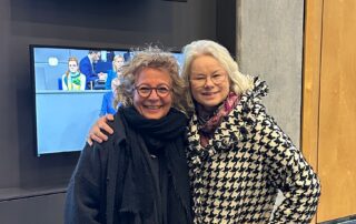 Beate Müller-Gemmeke und Kirsten Kappert-Gonther stehen nebeneinander und lächeln. Im Hintergrund ist ein Bildschirm mit einer Bundestagsszene zu sehen.