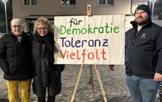 Drei Personen stehen abends auf einem Platz in Burladingen neben einem auf einer Staffelei befestigten Plakat mit der Aufschrift „für Demokratie, Toleranz, Vielfalt“. Rechts im Bild steht Maurice Rössler, Landtagskandidat. Neben Beate Müller-Gemmeke steht Annette Thriemer, eine der Initiator:innen der Mahnwache. Die Szene ist beleuchtet, winterlich gekleidet, ruhige Atmosphäre einer Mahnwache.