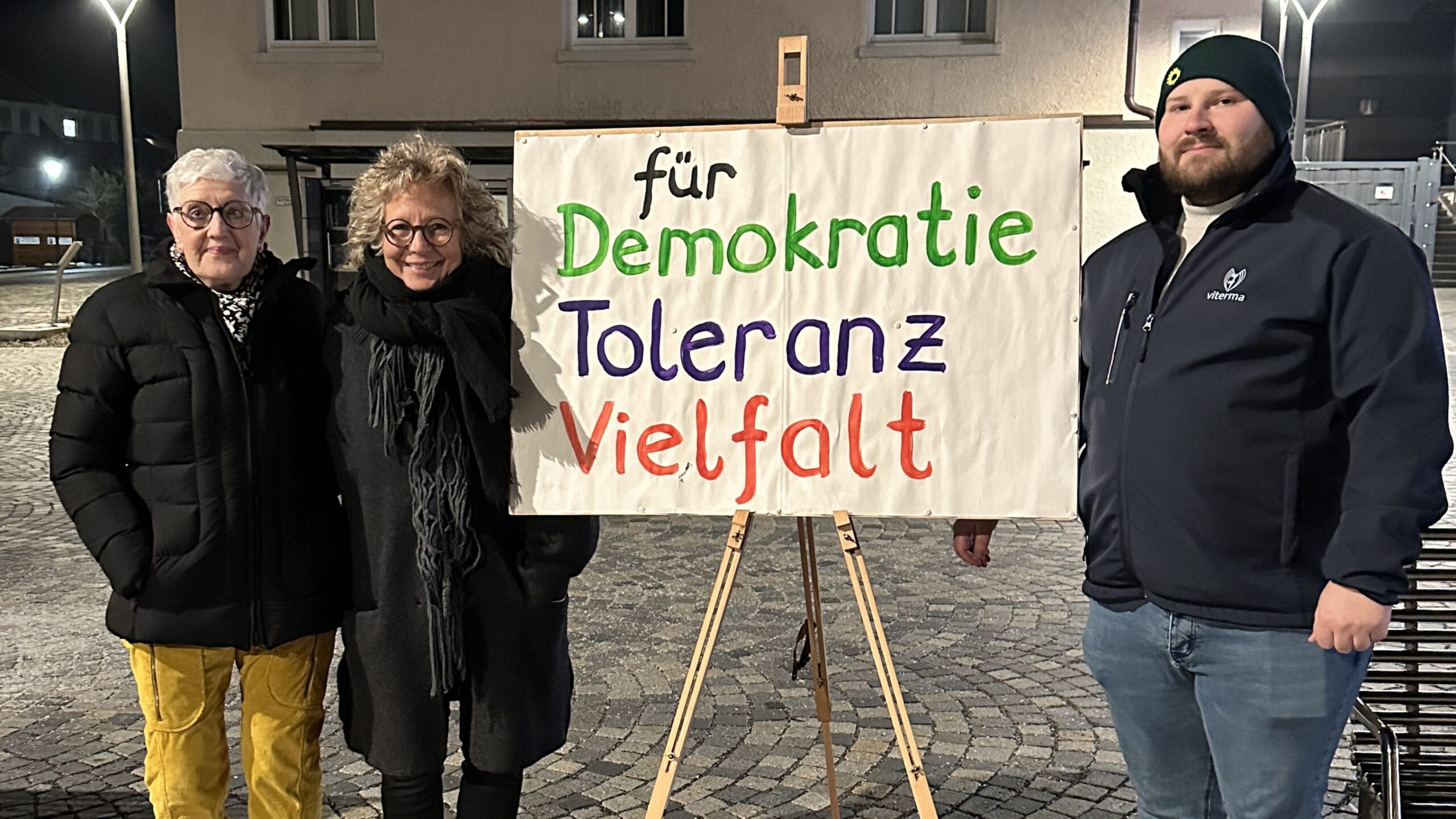 Drei Personen stehen abends auf einem Platz in Burladingen neben einem auf einer Staffelei befestigten Plakat mit der Aufschrift „für Demokratie, Toleranz, Vielfalt“. Rechts im Bild steht Maurice Rössler, Landtagskandidat. Neben Beate Müller-Gemmeke steht Annette Thriemer, eine der Initiator:innen der Mahnwache. Die Szene ist beleuchtet, winterlich gekleidet, ruhige Atmosphäre einer Mahnwache.