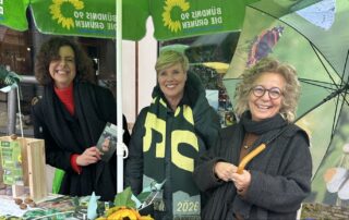 Lisa Weigert, Cindy Holmberg (Kandidatin) und Beate Müller-Gemmeke stehen am grünen Wahlkampf-Infostand von Bündnis 90/Die Grünen. Auf dem Tisch liegen Flyer und Infomaterial, vorne hängt ein Plakat mit Cem Özdemir. Eine große Sonnenblume ist am Stand befestigt. Die Stimmung ist offen und freundlich.