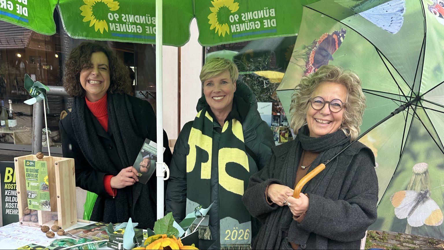 Lisa Weigert, Cindy Holmberg (Kandidatin) und Beate Müller-Gemmeke stehen am grünen Wahlkampf-Infostand von Bündnis 90/Die Grünen. Auf dem Tisch liegen Flyer und Infomaterial, vorne hängt ein Plakat mit Cem Özdemir. Eine große Sonnenblume ist am Stand befestigt. Die Stimmung ist offen und freundlich.