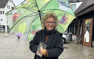 Beate Müller-Gemmeke steht beim Infostand in Metzingen bei Regen unter einem grünen Regenschirm mit Naturmotiven. Sie hält Wahlflyer in der Hand und lächelt in die Kamera.