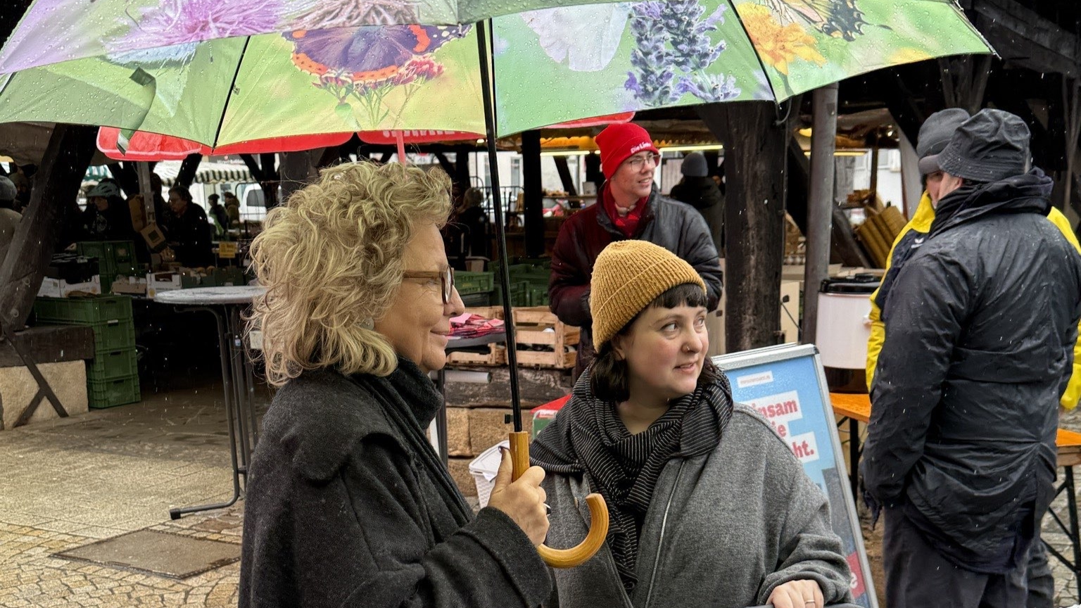 Beate Müller-Gemmeke spricht auf einem Wochenmarkt unter einem grünen Regenschirm mit einer jungen Frau mit Kinderwagen. Im Hintergrund sind Marktstände und weitere Menschen zu sehen. Die Szene zeigt ein persönliches Gespräch.