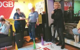 Beate Müller-Gemmeke spricht beim DGB-Format „Wahlopoly“ in Hechingen in ein Mikrofon. Sie steht auf einem großen Spielbrett mit markierten Politikfeldern wie Arbeitsmarkt, Kommunalfinanzen und Tarifbindung. Weitere Teilnehmende sitzen und stehen daneben, hören zu und verfolgen die Diskussion. Der Raum ist hell und lebendig