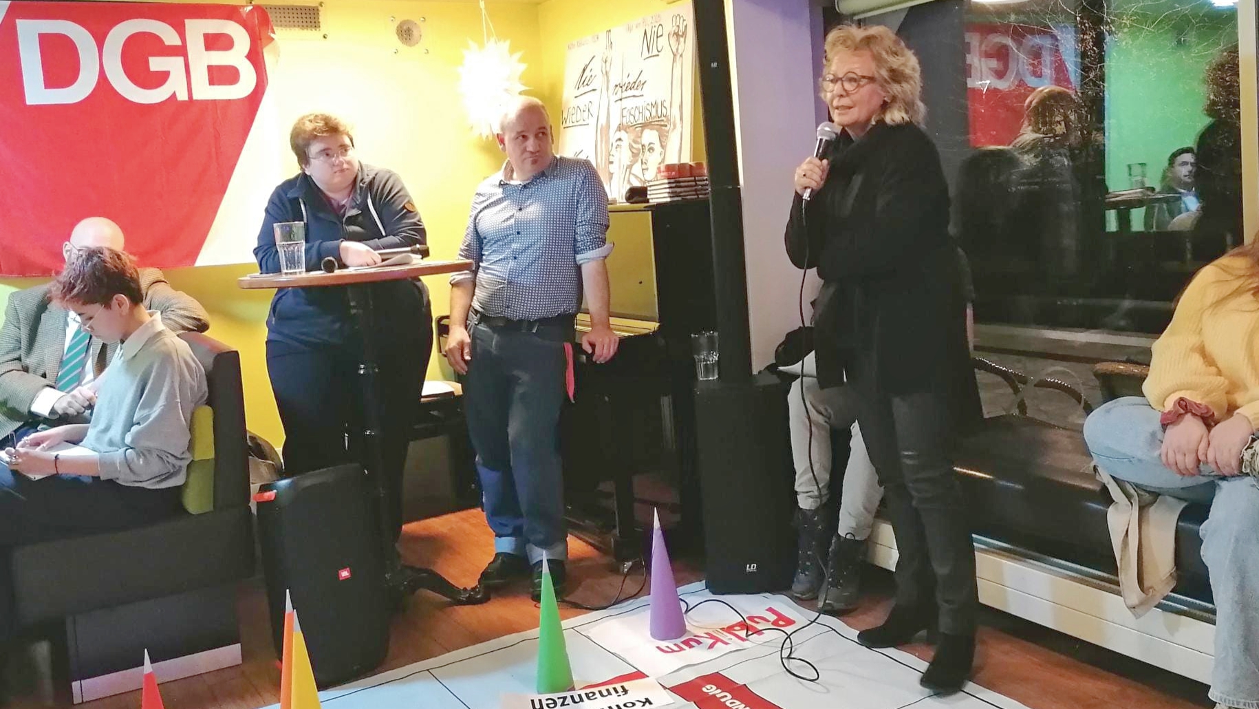 Beate Müller-Gemmeke spricht beim DGB-Format „Wahlopoly“ in Hechingen in ein Mikrofon. Sie steht auf einem großen Spielbrett mit markierten Politikfeldern wie Arbeitsmarkt, Kommunalfinanzen und Tarifbindung. Weitere Teilnehmende sitzen und stehen daneben, hören zu und verfolgen die Diskussion. Der Raum ist hell und lebendig