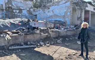 Beate Müller-Gemmeke steht vor den Trümmern eines zerstörten Schulgebäudes in der Ukraine. Die Außenwände sind eingestürzt, Ziegel und Schutt liegen auf dem Boden – ein sichtbares Zeichen der Zerstörung ziviler Infrastruktur.
