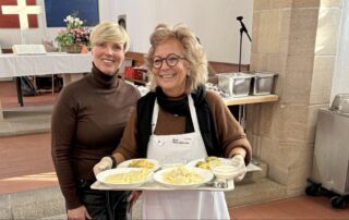 Cindy Holmberg steht neben Beate Müller-Gemmeke in der Vesperkirche Reutlingen. Beate trägt eine Schürze und hält ein Tablett mit Essen. Beide lächeln in die Kamera, im Hintergrund sind Tische und der Altarraum zu sehen.