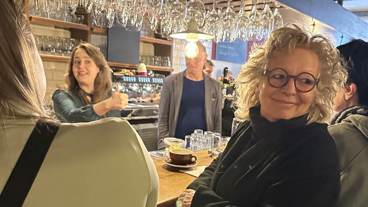 Beate Müller-Gemmeke steht an der Theke im Gespräch mit Gästen. Im Hintergrund ist Ricarda Lang zu sehen, die ebenfalls mit Besucher:innen spricht. Das Café ist voll, die Stimmung offen und kommunikativ.