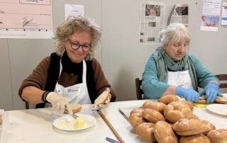 Beate Müller-Gemmeke und Hanne Krönke bereiten in der Vesperkirche Reutlingen Brötchen vor. Beide tragen Schürzen und Handschuhe, auf dem Tisch liegen Brote, Messer und Aufstrich.