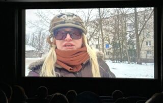 Filmszene im Kinosaal: Die Journalistin Zarina Zabrisky berichtet im Winter aus der ukrainischen Stadt Cherson; im Vordergrund sind die Silhouetten des Publikums zu erkennen.