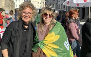 Beate Müller-Gemmeke steht auf einem Marktplatz neben einer jungen Frau, die eine grüne Fahne mit gelber Sonnenblume trägt. Beide lächeln in die Kamera. Im Hintergrund sind Menschen und Marktstände zu sehen.