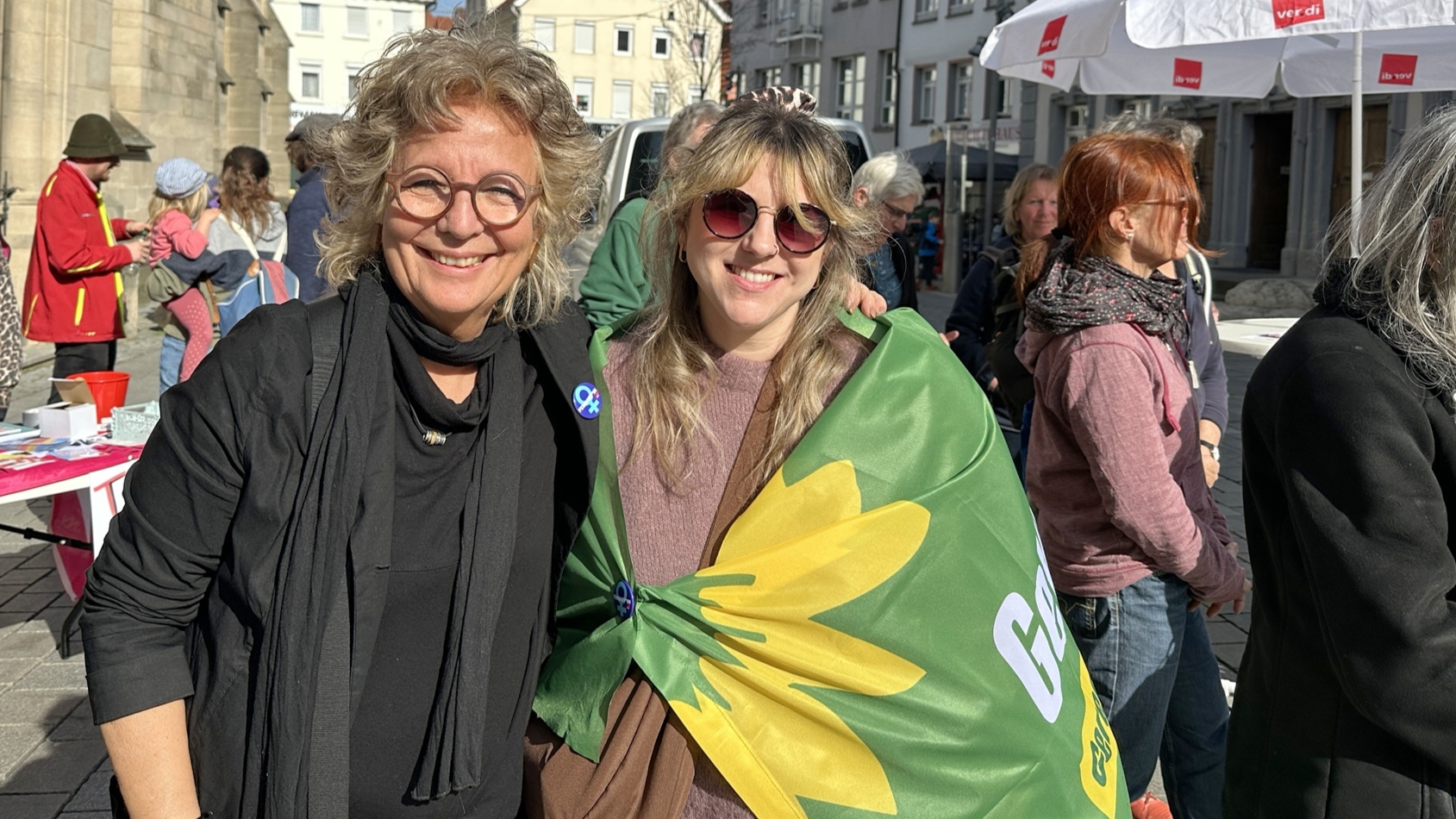 Beate Müller-Gemmeke steht auf einem Marktplatz neben einer jungen Frau, die eine grüne Fahne mit gelber Sonnenblume trägt. Beide lächeln in die Kamera. Im Hintergrund sind Menschen und Marktstände zu sehen.