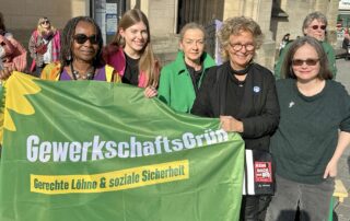 Mehrere Frauen stehen gemeinsam auf einem Platz vor einer Kirche und halten ein grünes Banner von GewerkschaftsGrün mit der Aufschrift „Gerechte Löhne & soziale Sicherheit“. Beate Müller-Gemmeke steht in der Mitte der Gruppe.