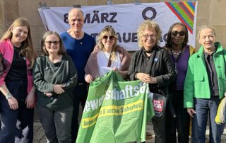 Gruppe von Frauen und Unterstützenden vor einem Banner zum Internationalen Frauentag am 8. März. In der Mitte steht Beate Müller-Gemmeke mit einer grünen Fahne von GewerkschaftsGrün. Die Gruppe posiert gemeinsam für ein Foto.