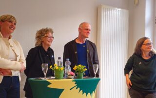 Beate Müller-Gemmeke steht zusammen mit mehreren Personen an einem Stehtisch bei einer Veranstaltung. Auf dem Tisch stehen Blumen, Gläser und Wasserflaschen. Die Gruppe hört aufmerksam zu und lächelt.