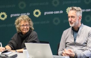 Beate Müller-Gemmeke und Volker Morbe im Gespräch während der Veranstaltung von GewerkschaftsGrün Saar. Beide sitzen nebeneinander an einem Tisch, vor ihnen ein Laptop und Notizen, im Hintergrund das grüne Logo der saarländischen Grünen.