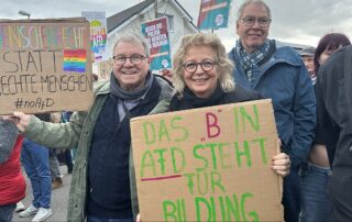 Beate Müller-Gemmeke steht mit Mitdemonstrierenden eng beieinander und hält ein selbstgemaltes Pappschild mit der Aufschrift „Das ‚B‘ in AfD steht für Bildung“. Daneben ist ein weiteres Schild zu sehen: „Menschenrechte statt rechte Menschen #noAfD“. Im Hintergrund weitere Banner gegen Faschismus und Rechtsextremismus.