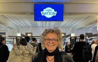 Ich stehe im Foyer des Friedrichstadtpalasts in Berlin und lächle in die Kamera. Über mir ist ein Bildschirm mit dem Logo „Lage der Nation“. Im Hintergrund stehen Besucher:innen der Live-Veranstaltung.