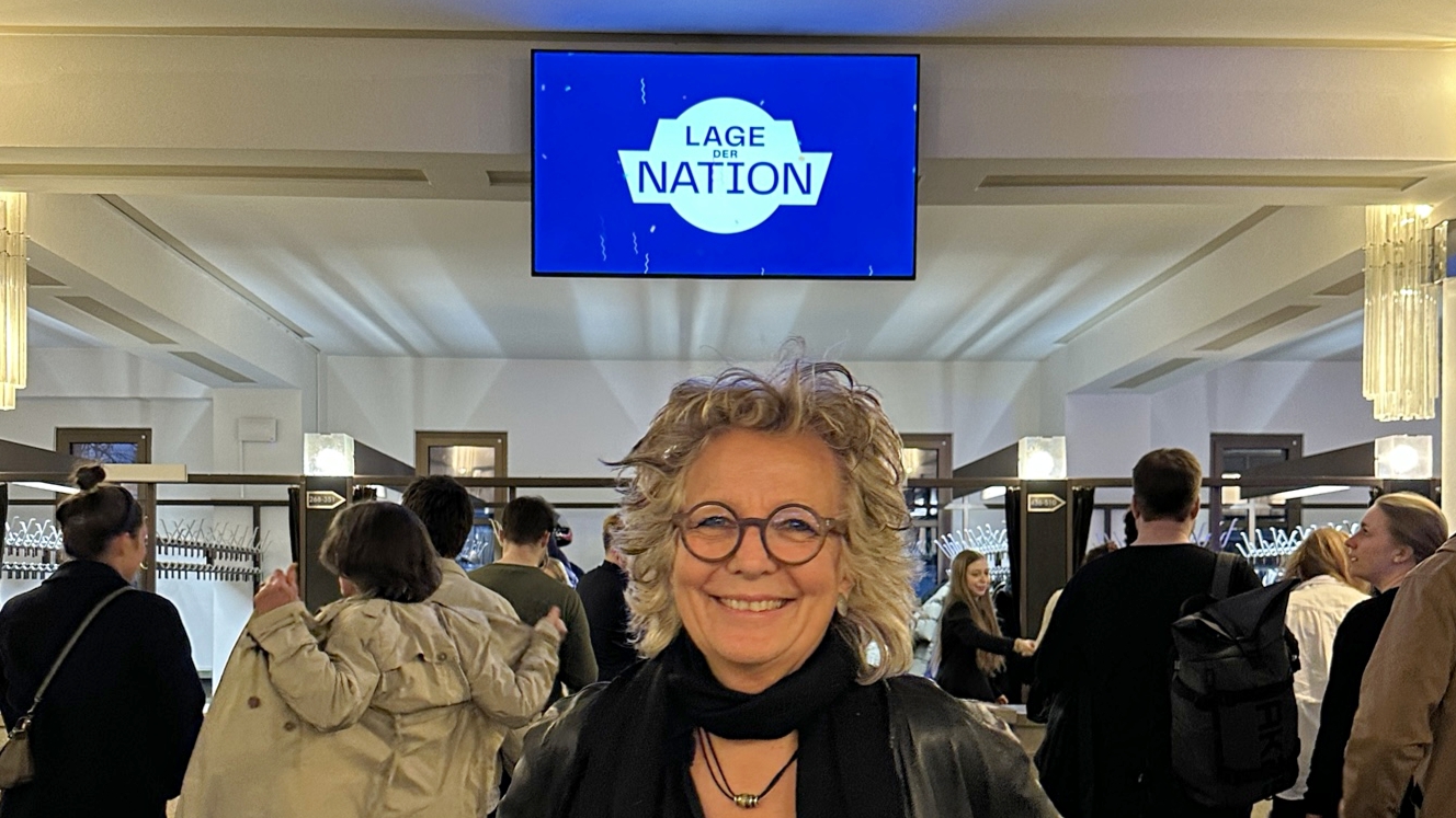 Ich stehe im Foyer des Friedrichstadtpalasts in Berlin und lächle in die Kamera. Über mir ist ein Bildschirm mit dem Logo „Lage der Nation“. Im Hintergrund stehen Besucher:innen der Live-Veranstaltung.