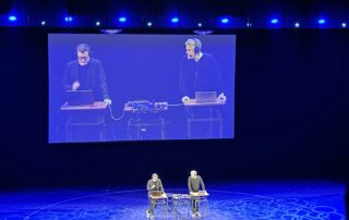 Bühne im Friedrichstadtpalast: Philip Banse und Ulf Buermeyer stehen an zwei Stehpulten mit Mikrofonen und Laptops und nehmen den Podcast „Lage der Nation“ live auf. Über ihnen ist eine große Leinwand mit Nahaufnahme.