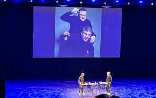 Große Bühne mit blauem Licht: Philip Banse und Ulf Buermeyer stehen an ihren Pulten. Auf der Leinwand darüber ist ein Foto der beiden zu sehen, in einer humorvollen Pose.