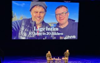 Bühne im Friedrichstadt-Palast: Philip Banse und Ulf Buermeyer stehen an ihren Pulten. Auf der großen Leinwand ist ein Foto von ihnen draußen zu sehen, mit der Aufschrift „Lage Intim – 10 Jahre in 20 Bildern“.