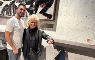 Beate Müller-Gemmeke steht gemeinsam mit Mojtaba Babaeifard in einem Innenraum vor einem großformatigen Kunstwerk. Beide lächeln in die Kamera. Mojtaba trägt ein weißes T-Shirt und Jeans, Beate eine schwarze Lederjacke, schwarzen Schal und runde Brille. Das Foto zeigt eine persönliche Begegnung im kulturellen Kontext.