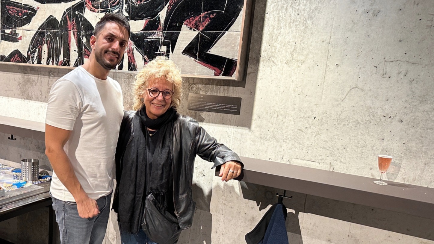 Beate Müller-Gemmeke steht gemeinsam mit Mojtaba Babaeifard in einem Innenraum vor einem großformatigen Kunstwerk. Beide lächeln in die Kamera. Mojtaba trägt ein weißes T-Shirt und Jeans, Beate eine schwarze Lederjacke, schwarzen Schal und runde Brille. Das Foto zeigt eine persönliche Begegnung im kulturellen Kontext.