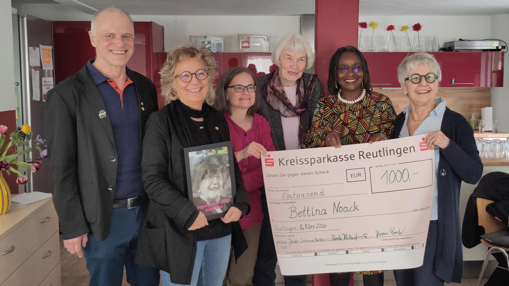 Gruppenfoto im Mütter- und Nachbarschaftszentrum Reutlingen bei der Verleihung des Irmgard-Zecher-Preises. Bettina Noack erhält die Auszeichnung; neben ihr stehen unter anderem Beate Müller-Gemmeke und weitere Unterstützer:innen. Gemeinsam halten sie einen symbolischen Scheck über 1.000 Euro, daneben ist ein gerahmtes Bild von Irmgard Zecher zu sehen.