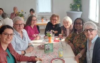 Bettina Noack und Beate Müller-Gemmeke sitzen gemeinsam mit anderen an einem Tisch im Mütter- und Nachbarschaftszentrum Reutlingen, trinken Kaffee und unterhalten sich. In der Mitte des Tisches steht ein kleines Bild von Irmgard Zecher. Die Atmosphäre wirkt herzlich und gemeinschaftlich.