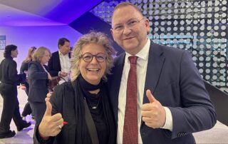 Du stehst lächelnd neben Frank Werneke, dem Bundesvorsitzenden von ver.di. Beide zeigen mit erhobenem Daumen in die Kamera. Im Hintergrund sind weitere Gäste der Jubiläumsfeier zu sehen.