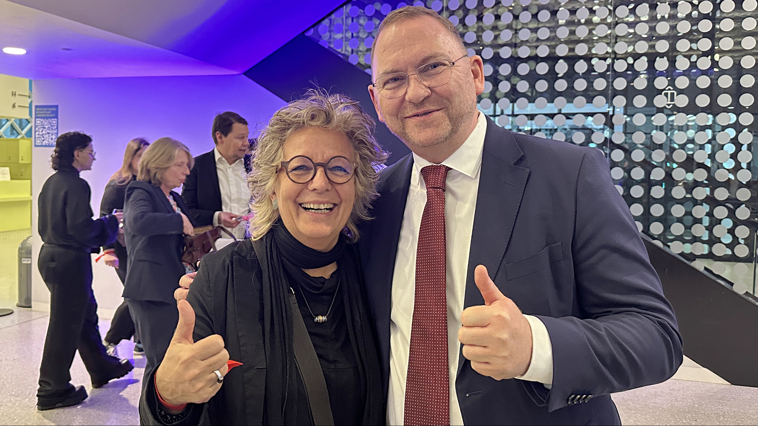 Du stehst lächelnd neben Frank Werneke, dem Bundesvorsitzenden von ver.di. Beide zeigen mit erhobenem Daumen in die Kamera. Im Hintergrund sind weitere Gäste der Jubiläumsfeier zu sehen.