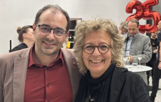 Du stehst neben Benjamin Stein, stellvertretender Landesbezirksleiter Baden-Württemberg. Im Hintergrund sind Gäste, Stehtische und rote Luftballons mit der Zahl „25“.