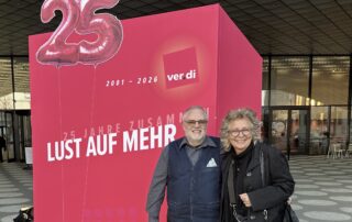 Du stehst neben Bernd Meffert, Beisitzer bei GewerkschaftsGrün, vor einer großen roten Wand mit der Aufschrift „25 Jahre ver.di – Lust auf mehr“. Darüber sind Luftballons in Form der Zahl 25 zu sehen.