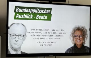 Ein großer Bildschirm zeigt eine Präsentationsfolie mit der Überschrift „Bundespolitischer Ausblick – Beate“. Links ist ein Schwarz-Weiß-Porträt von Friedrich Merz, rechts ein farbiges Porträt von Beate Müller-Gemmeke. In der Mitte steht ein Zitat: „Der Sozialstaat, wie wir ihn heute haben, ist mit dem, was wir volkswirtschaftlich leisten, nicht mehr finanzierbar“ – Friedrich Merz, 23.08.2025.