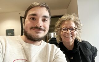 „Selfie von Beate Müller-Gemmeke (rechts) mit einem jungen Mann mit Bart und Brille. Beide lächeln in die Kamera. Der junge Mann trägt ein helles Sweatshirt, Beate Müller-Gemmeke eine schwarze Jacke. Im Hintergrund sind ein Flur und ein Hinweisschild zu erkennen.“