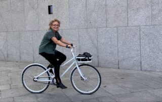 „Beate Müller-Gemmeke sitzt lächelnd auf einem weißen Fahrrad vor einer grauen Steinwand. Sie trägt ein grünes Oberteil, schwarze Hose und schwarze Schuhe. Das Fahrrad hat einen Gepäckträger mit einer schwarzen Tasche. Die Szene wirkt urban und fröhlich.“