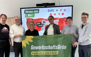 Sechs Personen stehen nebeneinander in einem Raum und halten gemeinsam ein grünes Banner mit der Aufschrift „GewerkschaftsGrün – Gerechte Löhne & soziale Sicherheit“. In der Mitte steht Beate Müller-Gemmeke, rechts neben ihr Finn Schwarz. Im Hintergrund ist ein Bildschirm mit der Aufschrift „Heraus zum 1. Mai! 1. Mai 2026“ sowie Logos von Gewerkschaften zu sehen. Die Gruppe lächelt in die Kamera.
