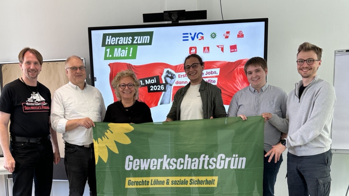 Sechs Personen stehen nebeneinander in einem Raum und halten gemeinsam ein grünes Banner mit der Aufschrift „GewerkschaftsGrün – Gerechte Löhne & soziale Sicherheit“. In der Mitte steht Beate Müller-Gemmeke, rechts neben ihr Finn Schwarz. Im Hintergrund ist ein Bildschirm mit der Aufschrift „Heraus zum 1. Mai! 1. Mai 2026“ sowie Logos von Gewerkschaften zu sehen. Die Gruppe lächelt in die Kamera.