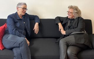 Bild 4: „Beate Müller-Gemmeke (rechts) im Gespräch mit Sylvia Rietenberg (links). Beide sitzen entspannt auf einem schwarzen Sofa in einem hellen Raum. Sylvia Rietenberg trägt ein dunkles Oberteil, Beate Müller-Gemmeke eine schwarze Jacke über einem dunklen Pullover. Im Hintergrund sind Türen und ein rotes Kissen zu sehen.“