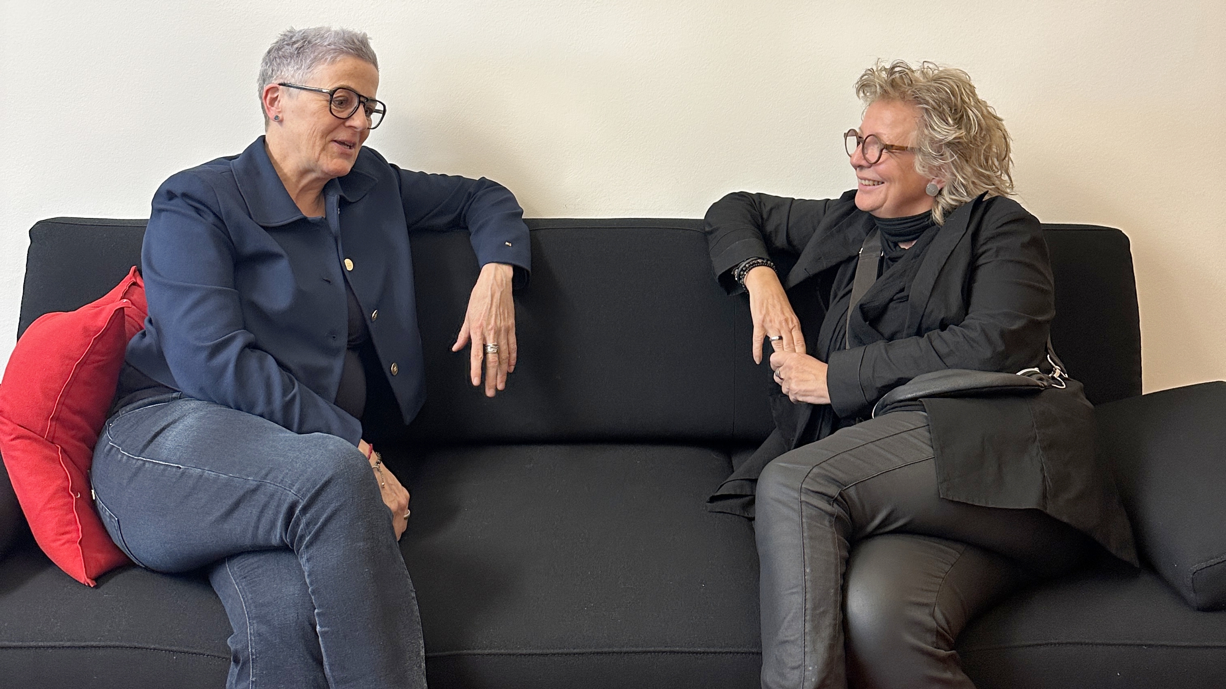 Bild 4: „Beate Müller-Gemmeke (rechts) im Gespräch mit Sylvia Rietenberg (links). Beide sitzen entspannt auf einem schwarzen Sofa in einem hellen Raum. Sylvia Rietenberg trägt ein dunkles Oberteil, Beate Müller-Gemmeke eine schwarze Jacke über einem dunklen Pullover. Im Hintergrund sind Türen und ein rotes Kissen zu sehen.“