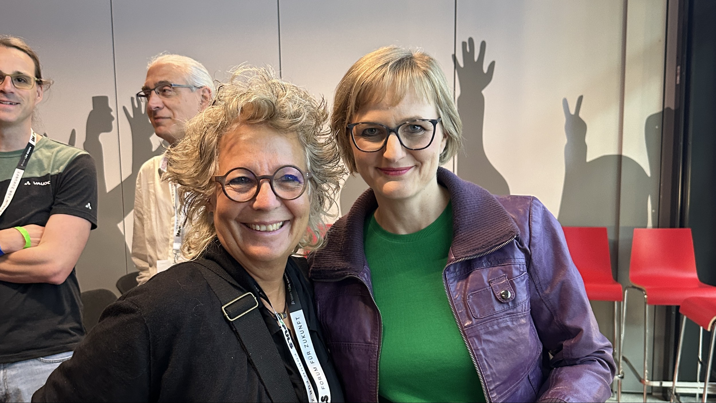 Beate Müller-Gemmeke und Franziska Brantner, Bundesvorsitzende von Bündnis 90/Die Grünen, stehen beim IM/PULS-Forum nebeneinander und lächeln in die Kamera. Im Hintergrund sind rote Stühle und stilisierte Hand-Schatten an einer Wand zu sehen.