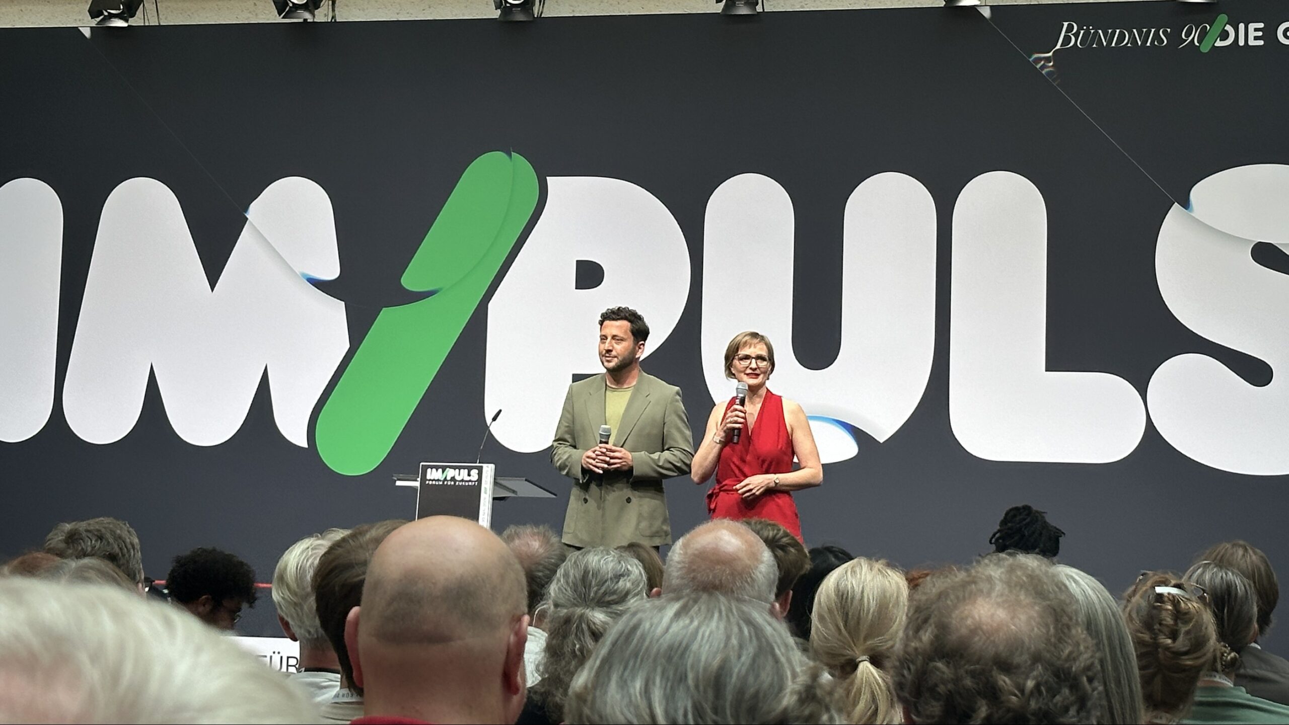 Franziska Brantner und Felix Banaszak eröffnen das IM/PULS-Forum auf der Bühne vor dem großen Veranstaltungslogo. Im Vordergrund sind die Köpfe zahlreicher Zuhörer*innen zu sehen.