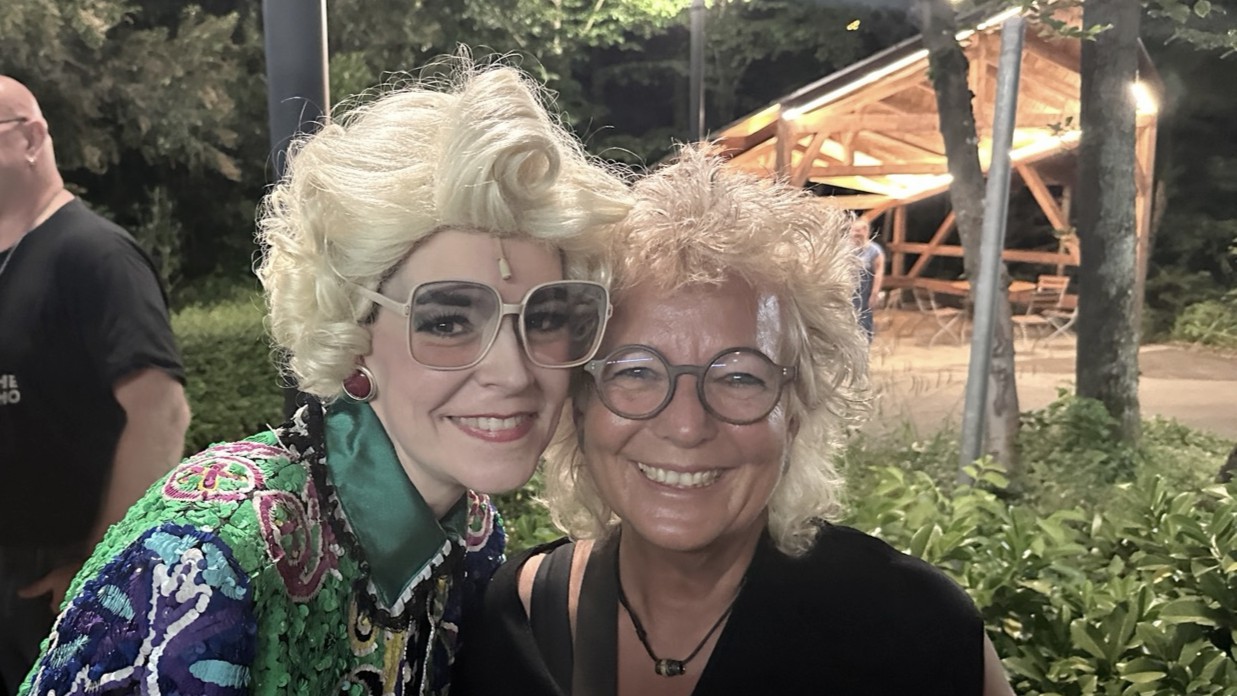 Selfie im Naturtheater Reutlingen: Beate Müller-Gemmeke steht lächelnd neben der Darstellerin der Claire Zachanassian, die mit blonder Perücke, großer Sonnenbrille und auffälligem, bunt glitzerndem Kostüm zu sehen ist. Im Hintergrund sind Grünflächen und Holzbauten des Naturtheaters im Abendlicht zu erkennen.